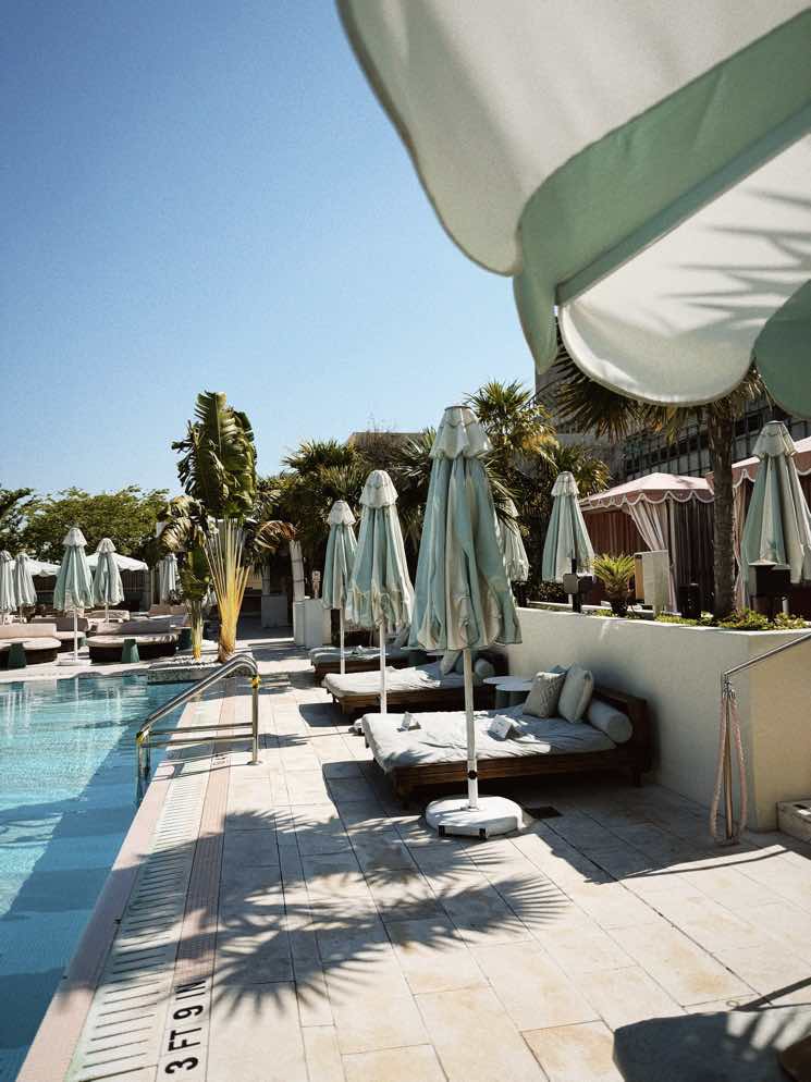 A sunlit poolside resort with pale green umbrellas, palm trees, and lounge cabanas